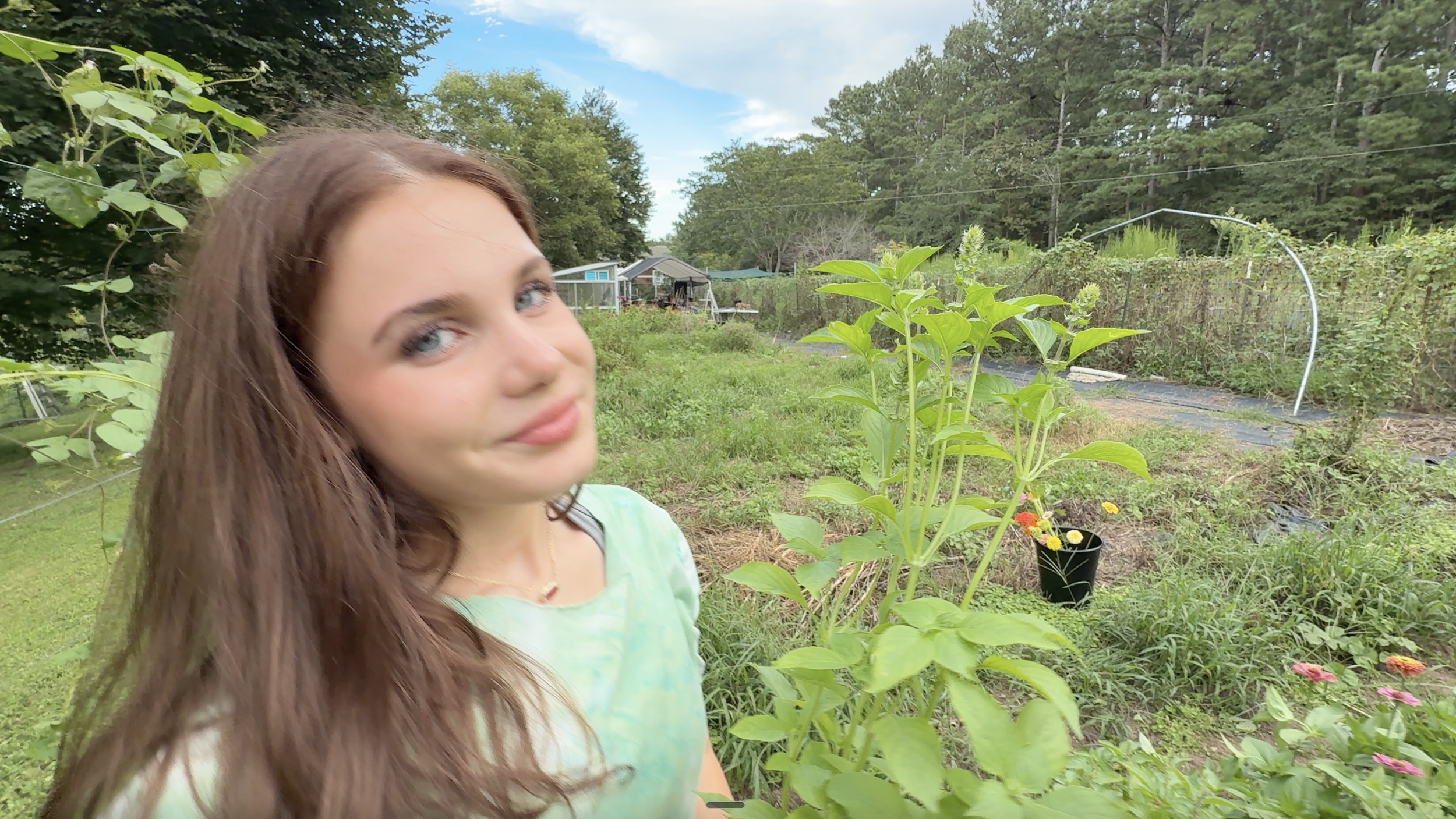 Young woman with long brown hair and blue eyes standing in a garden with green plants and trees, holding a plant, with a background of garden structures and a blue sky.
