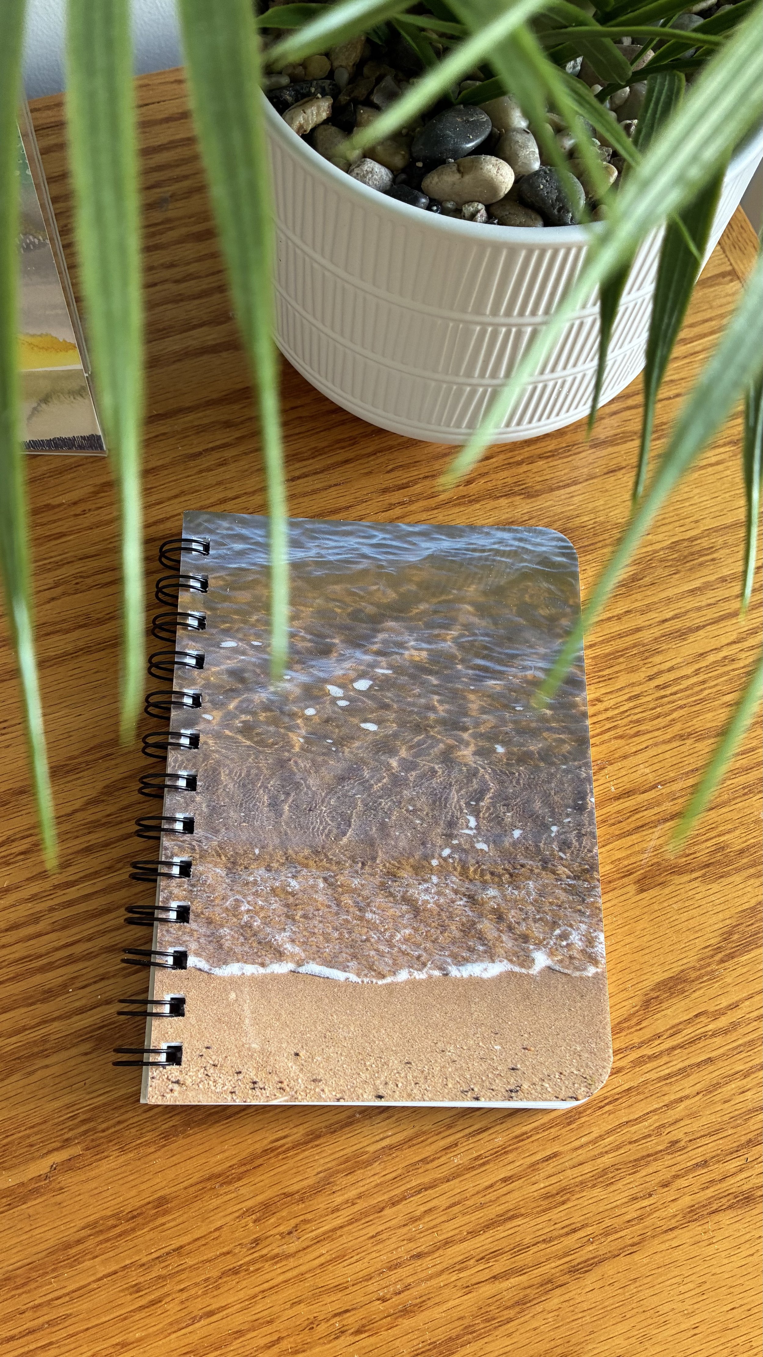 Top view of a notebook with ocean waves on its cover, resting on a wooden table. Behind it, there is a white pot with smooth stones and green plant leaves, partially obscuring the scene.