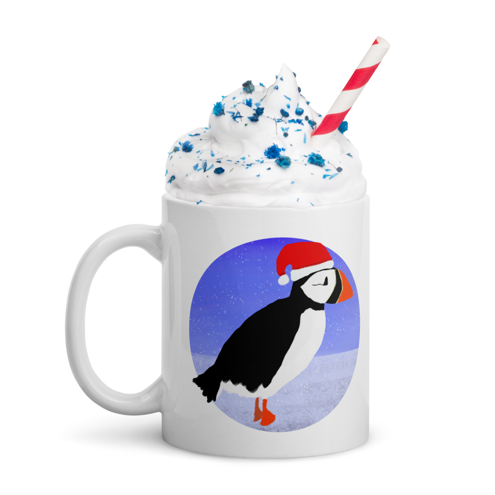 A white mug with a penguin wearing a Santa hat, standing on snow with a purple and blue sky background. The mug is filled with whipped cream, sprinkled with blue confetti, and topped with a red and white striped straw. Bright Blue Art Company
