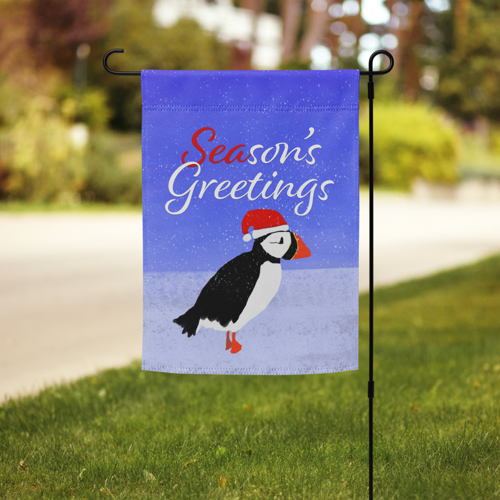 A garden flag with a penguin wearing a Santa hat, set against a snowy background, with the words "Season's Greetings" in red and white text. Holiday Shopping Bright Blue Art Company
