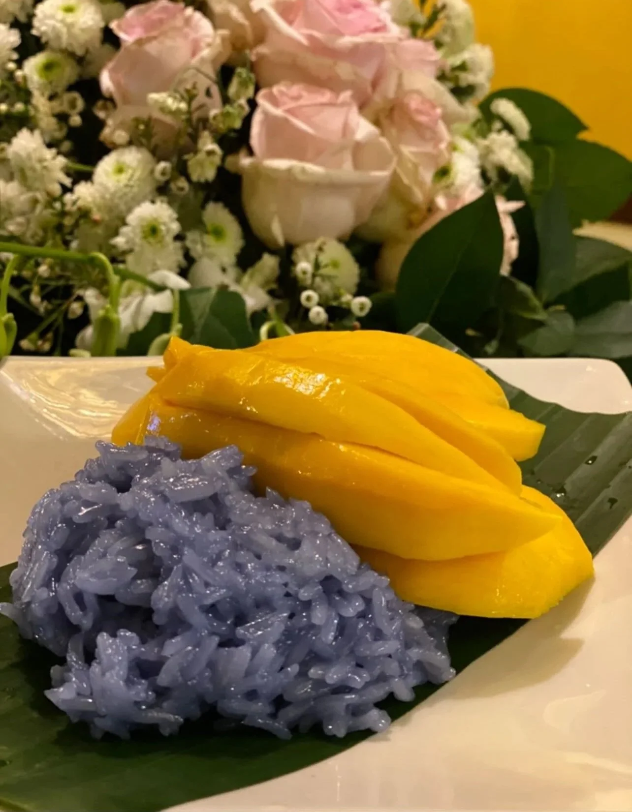 Purple sticky rice served with sliced mango on a white plate with green leaves, with a blurred background of pink and white flowers.