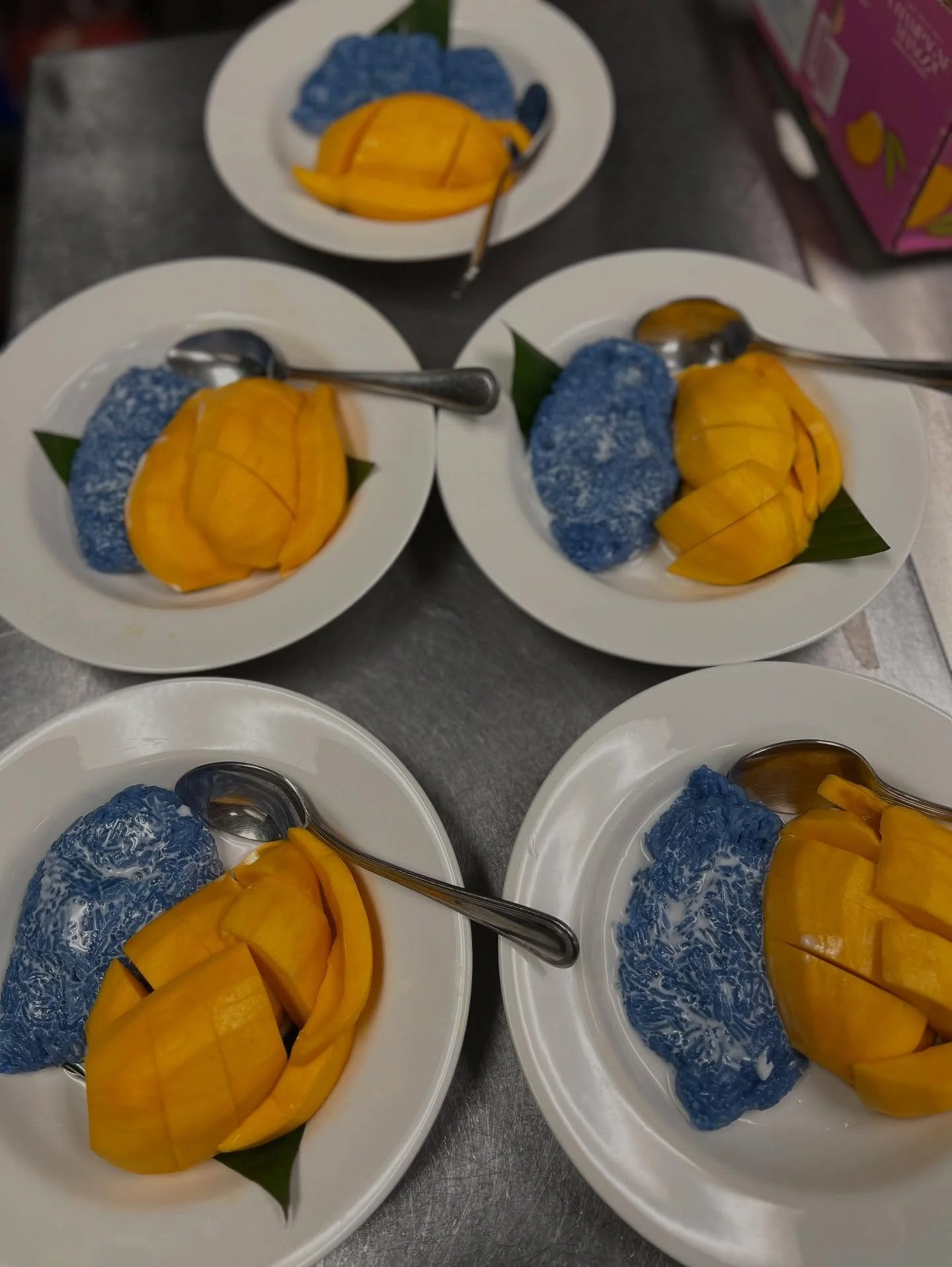 Four white plates with slices of ripe yellow mango and blue sticky rice topped with shredded coconut, each with a metal spoon, arranged on a metal surface.