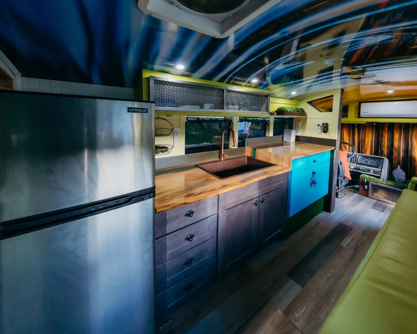 Compact kitchen with stainless steel refrigerator, wooden countertop, blue cabinet, and copper sink, inside a colorful camper van with a yellow sofa and a wood-paneled wall.