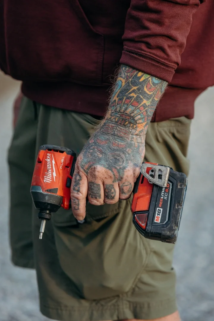 Close-up of a tattooed man holding a red Milwaukee cordless drill with a battery pack in his right hand, wearing a maroon hoodie and green shorts.