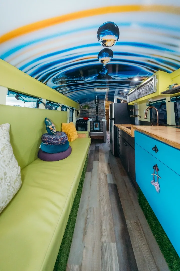 Interior of a tiny home with a colorful mural of a water droplet and ripples on the ceiling, featuring a lime green seating area with pillows, and a kitchen with wooden countertops, blue cabinets, and stainless steel appliances.