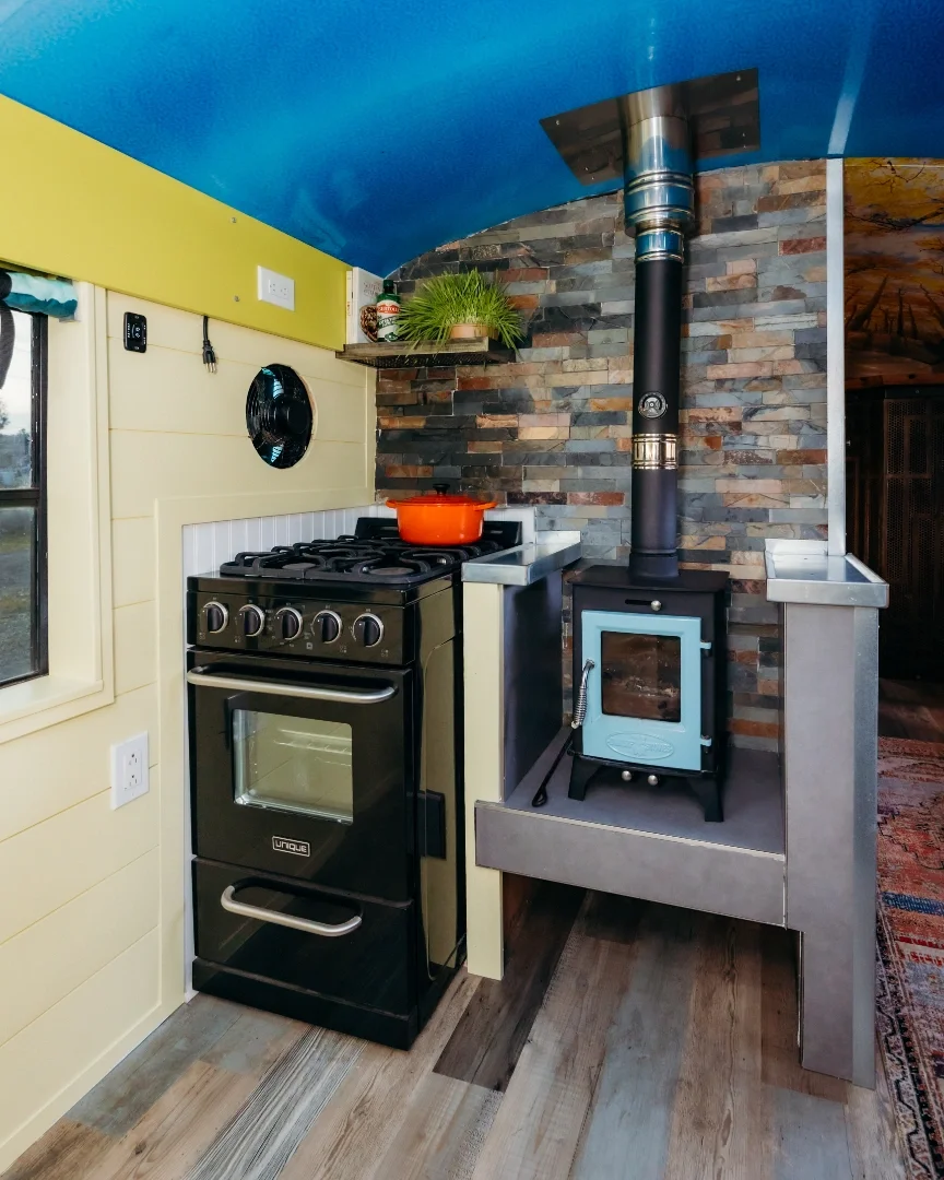 Small kitchen with a black stove next to a wood-burning stove with a chimney pipe on a raised platform, brick wall background, yellow wall, and wooden flooring.