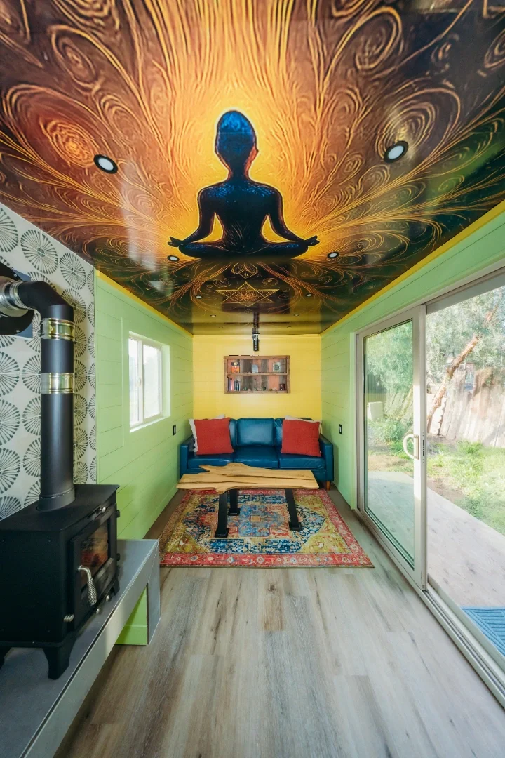 Room with a colorful ceiling mural of a person in a meditative pose surrounded by swirling patterns, yellow walls, a sliding glass door, a blue sofa with red pillows, a small wooden table, a patterned rug, and a wood stove.