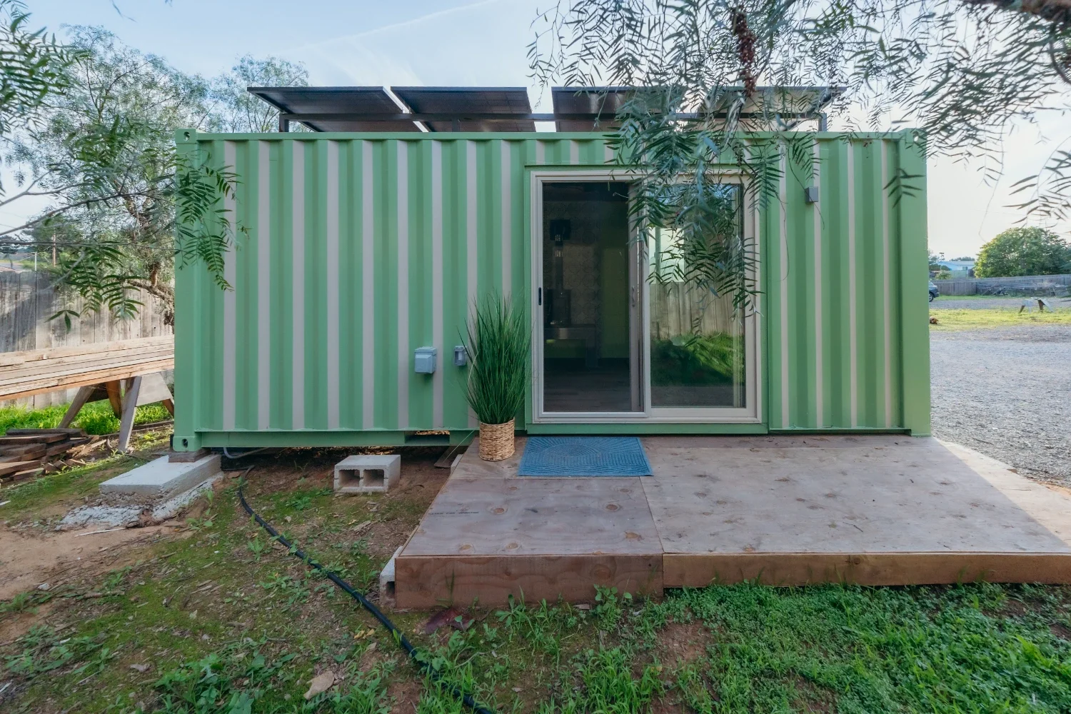 Green corrugated metal tiny house with sliding glass door, solar panels on roof, small wooden porch with a potted plant and blue mat, outdoor yard