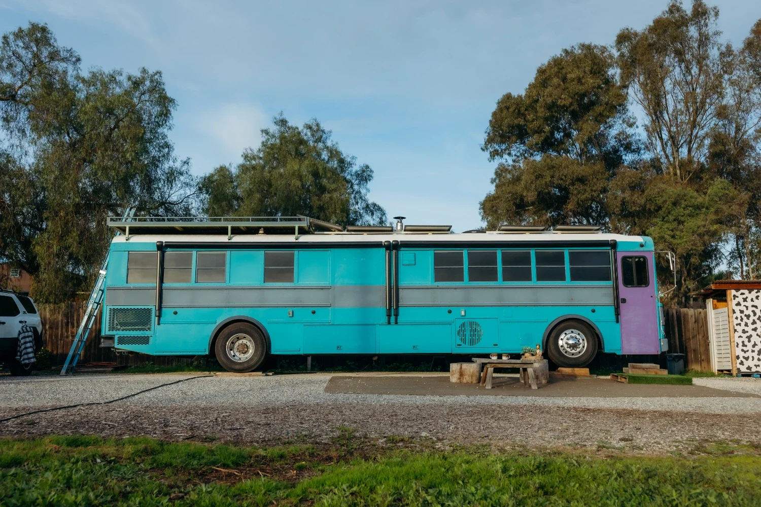 A colorful converted bus painted in blue with purple on the front, parked outdoors on gravel with trees in the background.