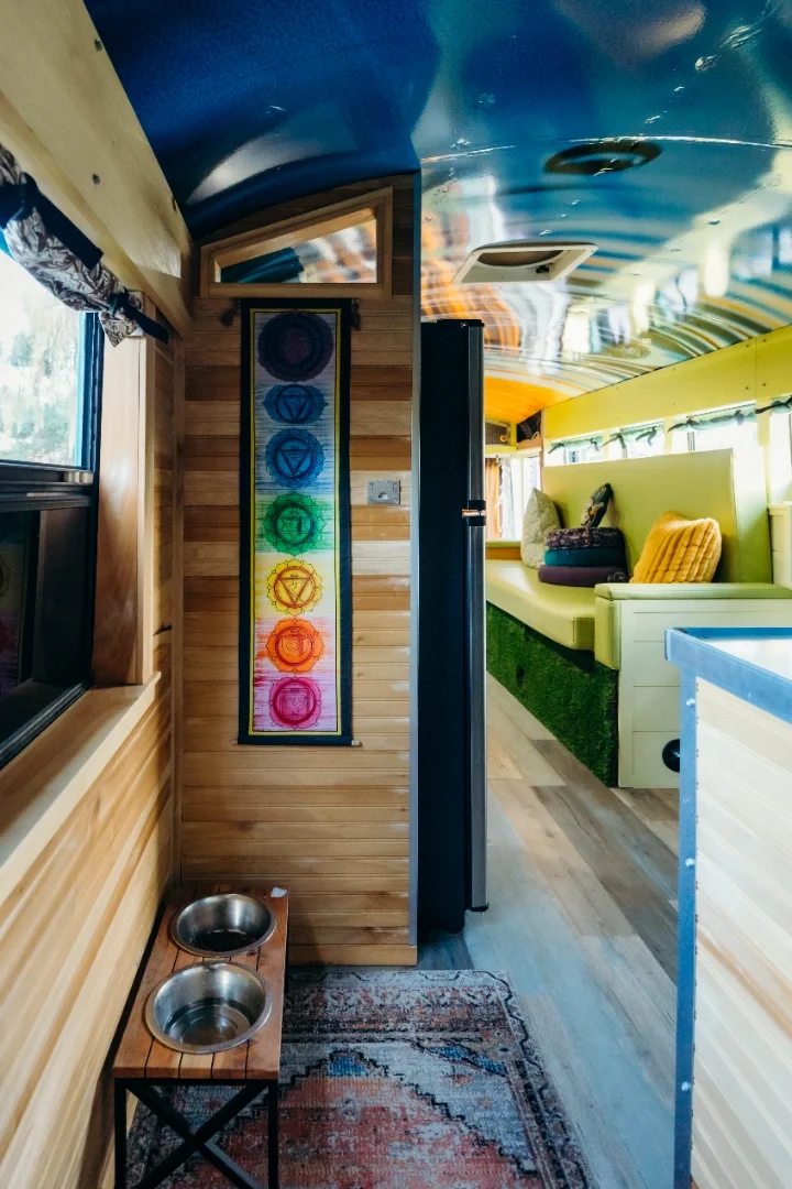 Interior of a cozy camper with wooden walls, a rainbow chakra art hanging, a small table with two metal pet bowls, and a yellow cushioned bench with pillows, illuminated by natural light from windows.