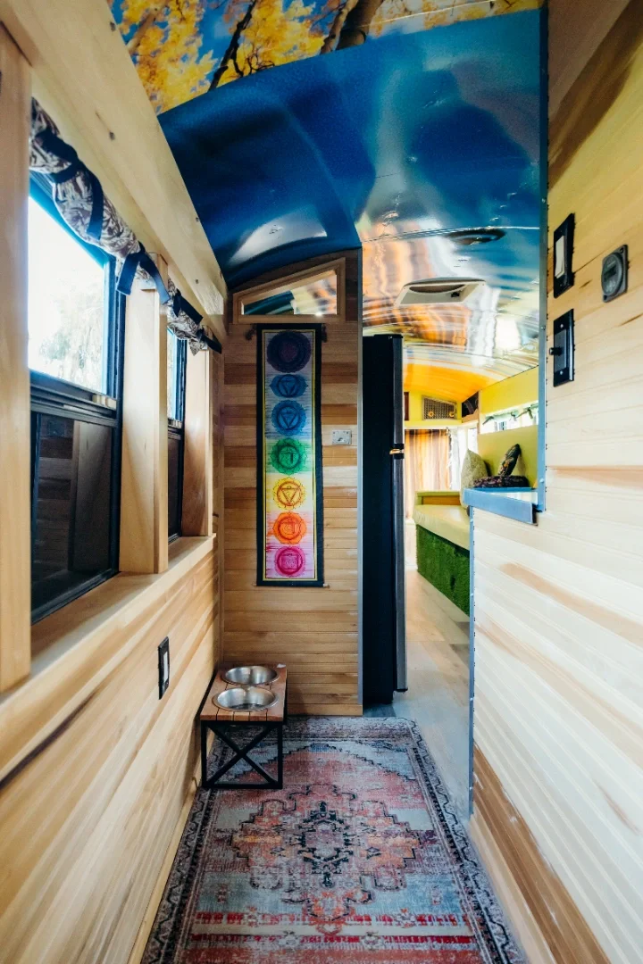 Narrow hallway inside a wooden tiny house with wood-paneled walls, a colorful Tibetan chakra painting on the wall, a small table with pet bowls, a patterned rug, windows with curtains, and a peek into a living area with a green couch.