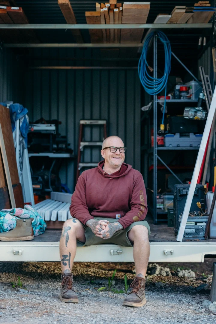 A man with glasses and tattoos on his legs is sitting outside on the edge of a small storage trailer, smiling. Behind him are shelves with tools and a blue coiled hose, in a garage or workshop setting.