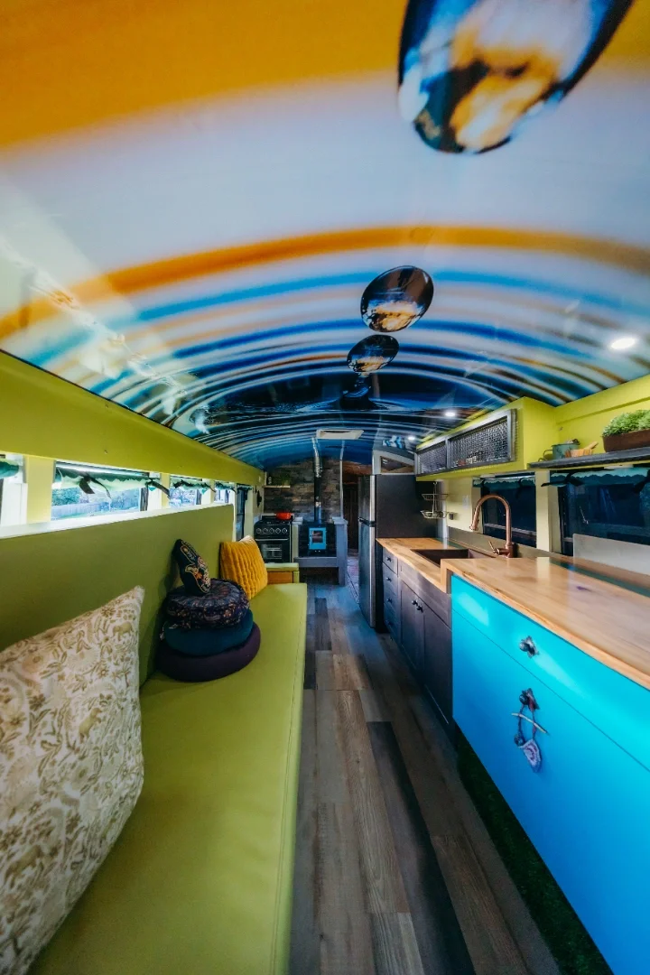 Interior of a camper van with a ceiling featuring a large photo print of water droplets rippling on a surface, yellow walls, green seating area with pillows, wooden kitchenette counter with a sink, and various appliances.