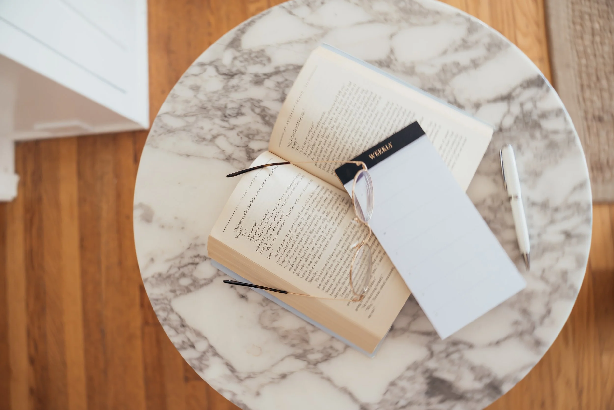 Open book with reading glasses and a notebook on a marble tabletop.