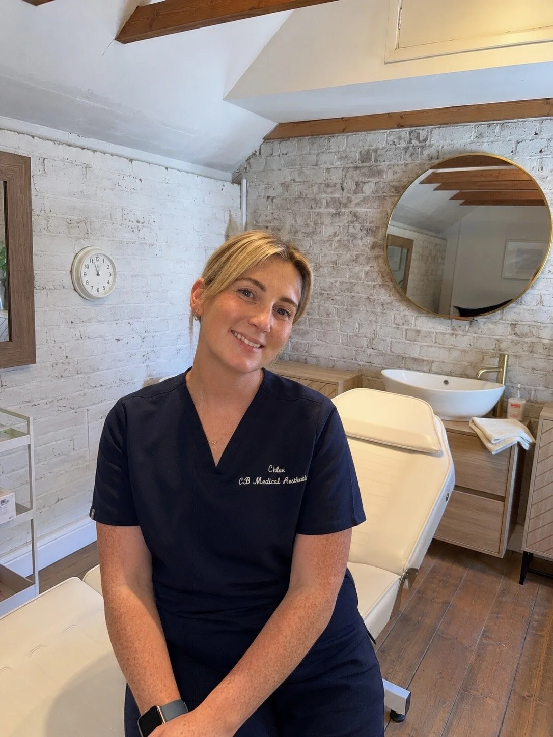 A woman with short blonde hair in medical scrubs sitting in a treatment room with a white brick wall, round mirror, and wooden accents. She is smiling at the camera.