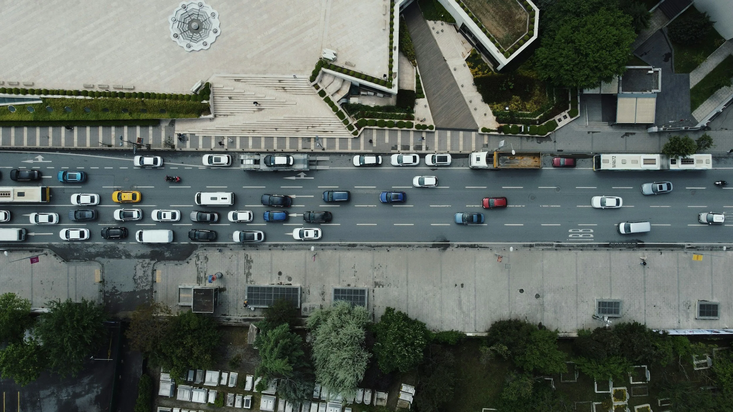 An aerial view of a busy city street with multiple cars, including a yellow taxi, and surrounding modern buildings and greenery.