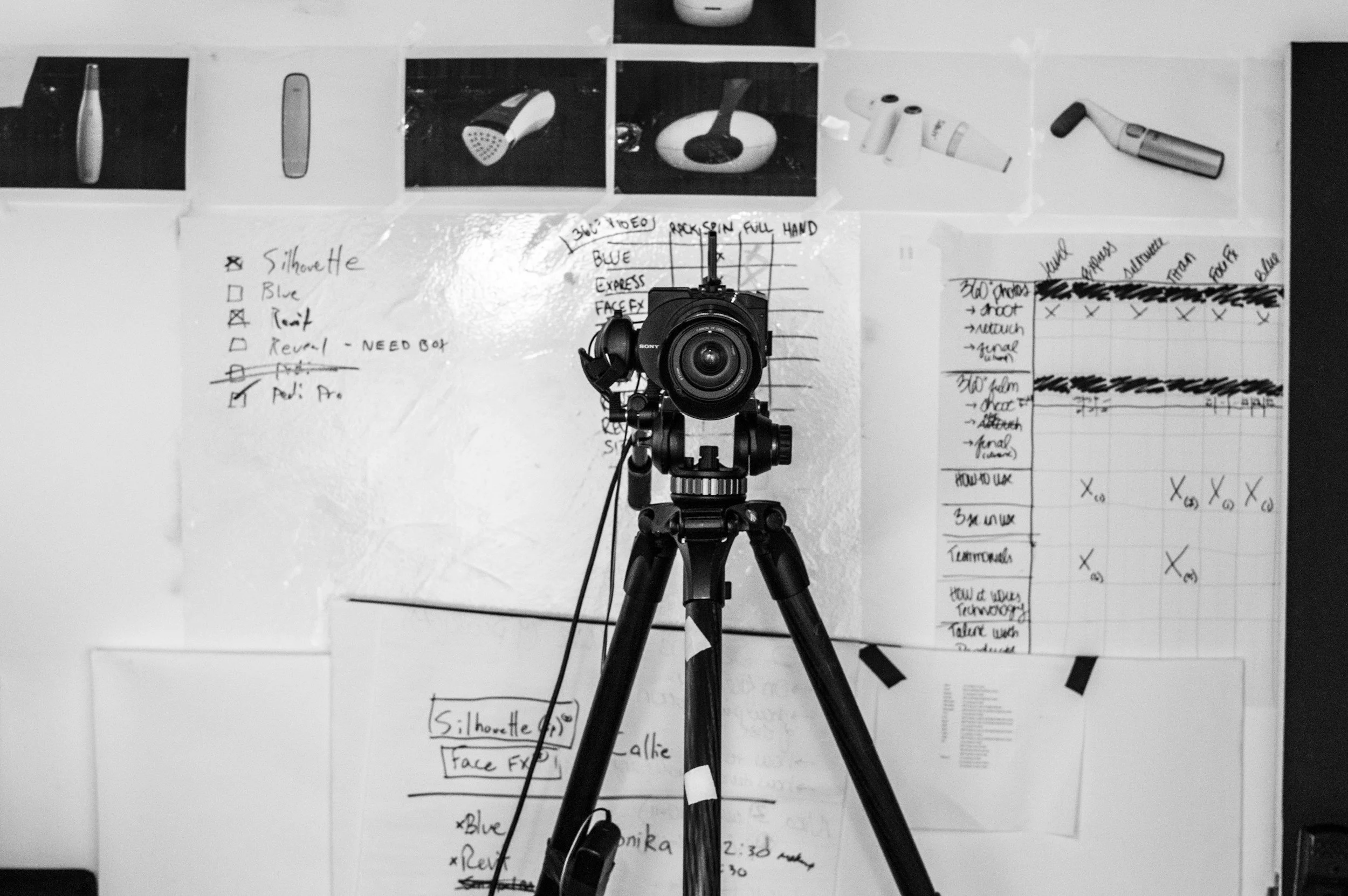 A black and white photo of a camera on a tripod positioned in front of a wall with various notes, charts, and photographs of different objects, including a hairbrush, a hairdryer, and a microphone, along with handwritten lists and diagrams.