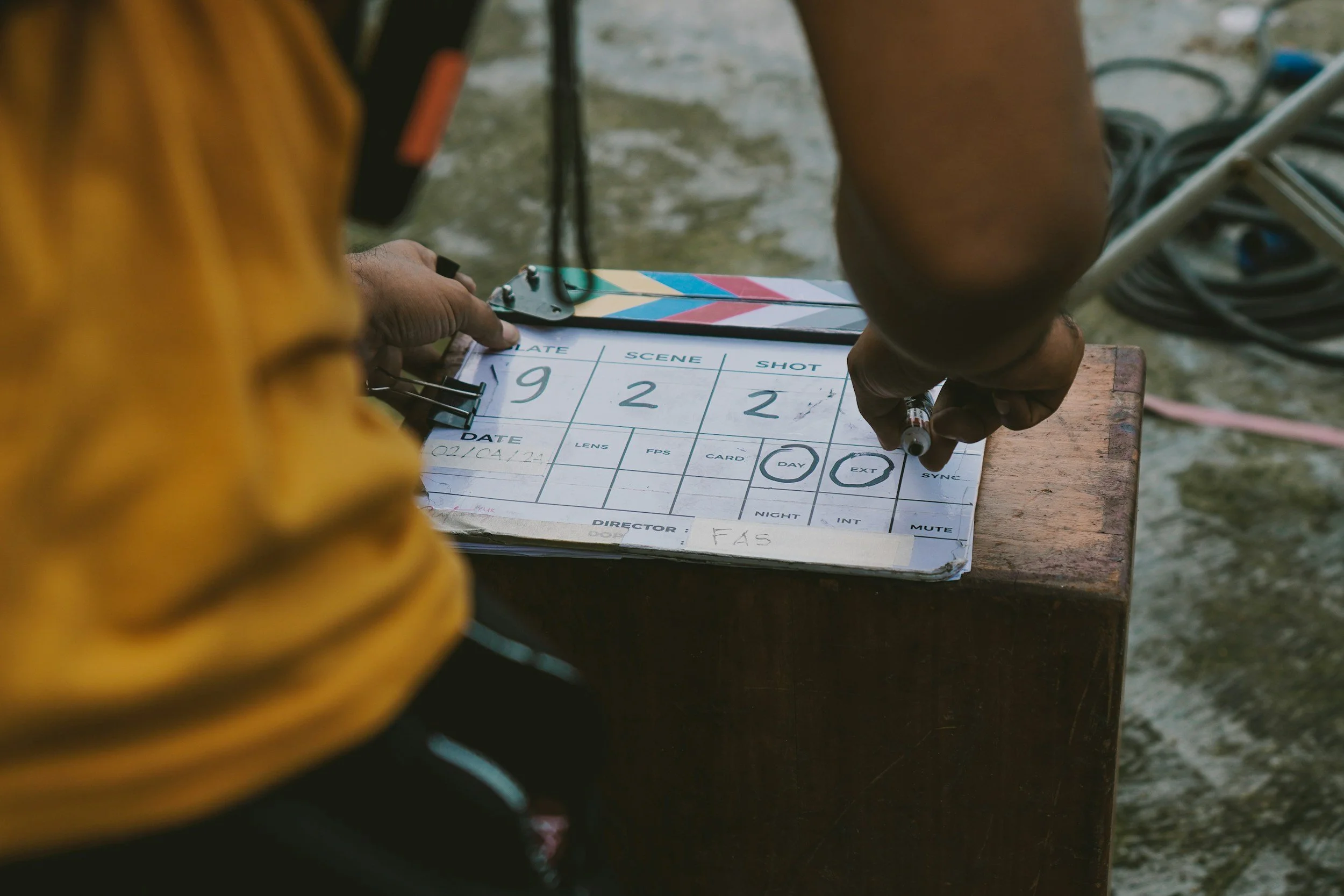 A person with a yellow shirt handles a clapperboard on a wooden surface near a beach, with coiled cables and equipment visible in the background.