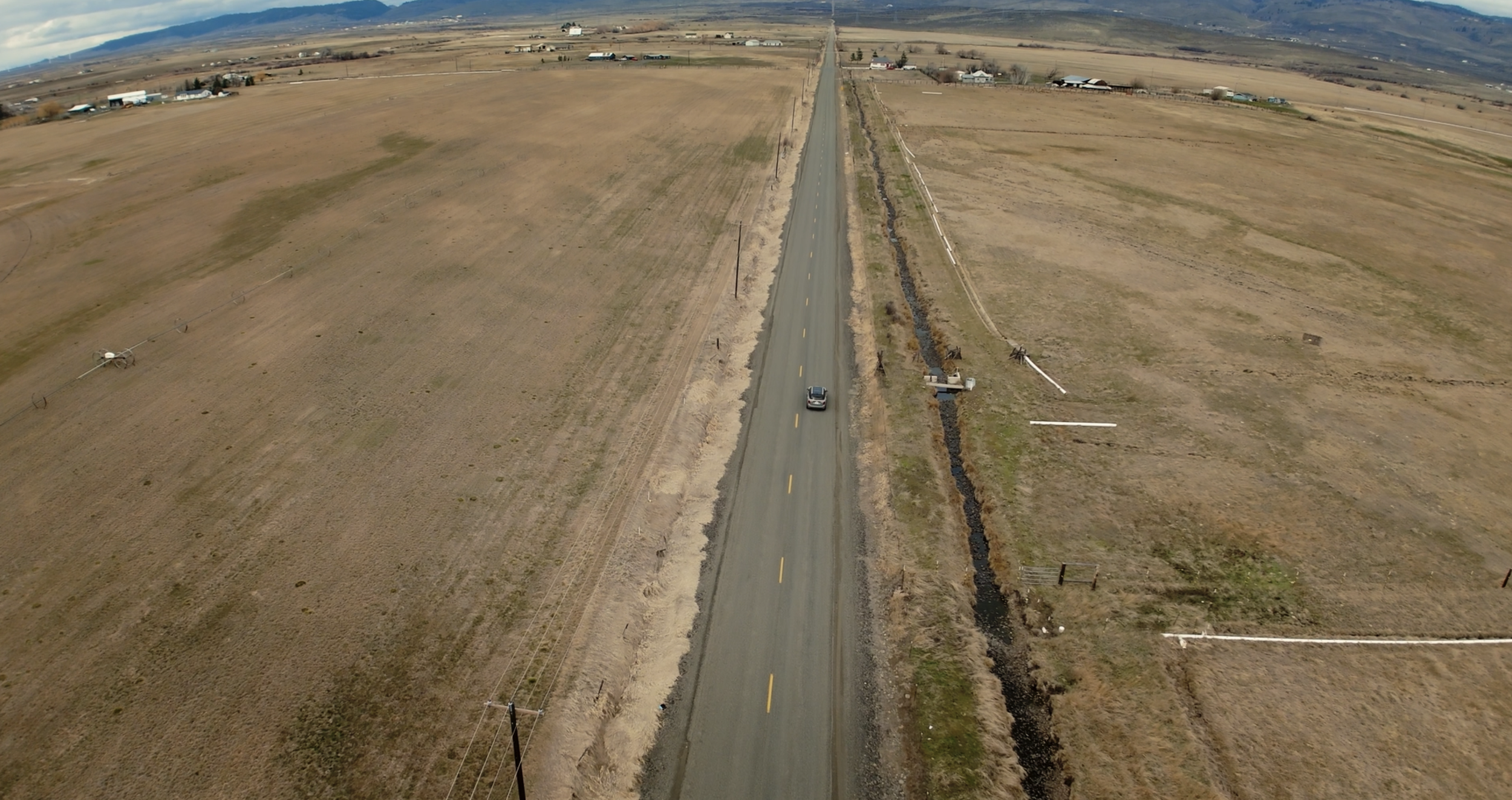 A long straight rural road running through open farmland with a single car driving along it. The landscape is mostly flat with some distant buildings and mountains on the horizon. There are utility poles along the road and a small drainage ditch on the right side.