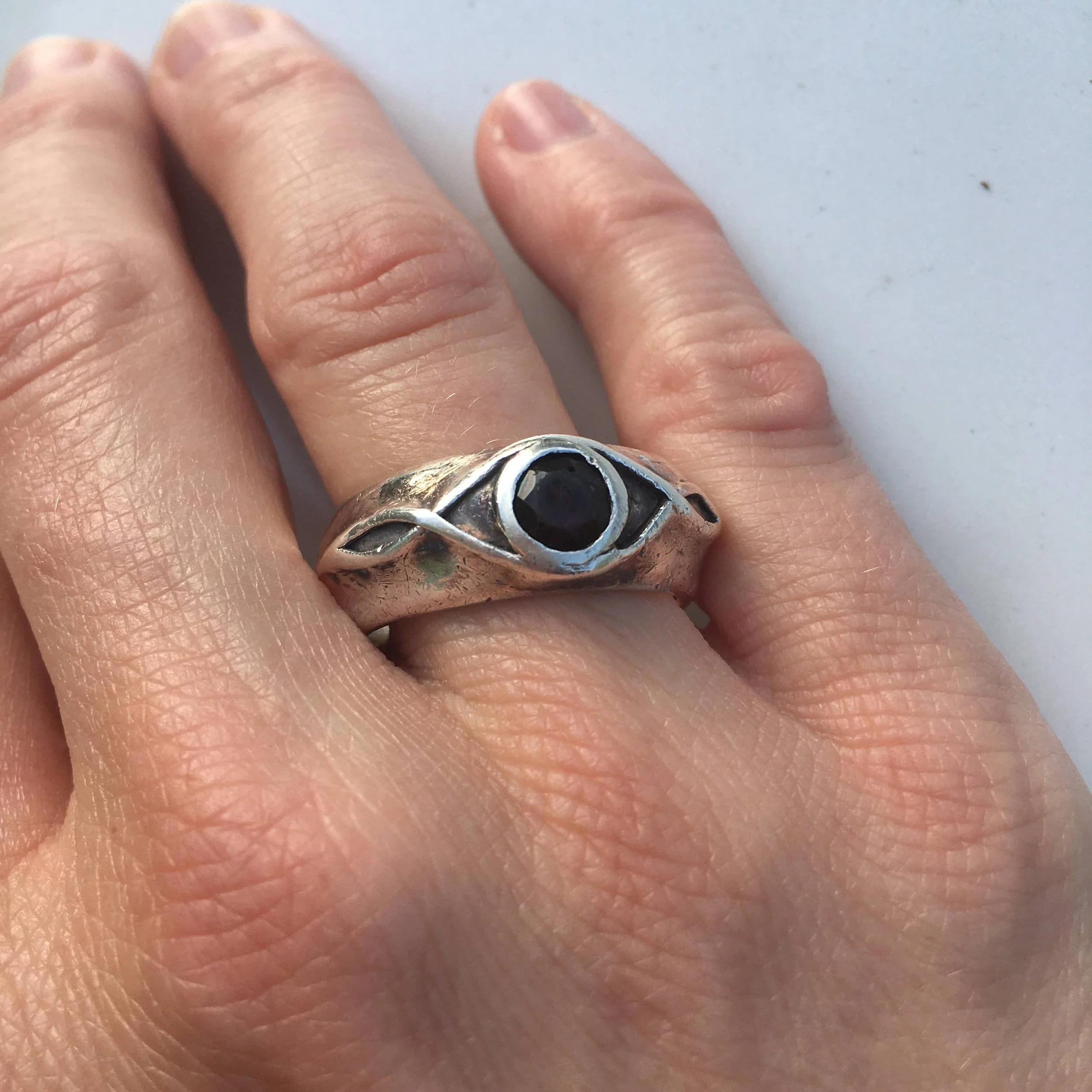 A person's finger wearing a silver ring with an eye design and a black gemstone in the center.