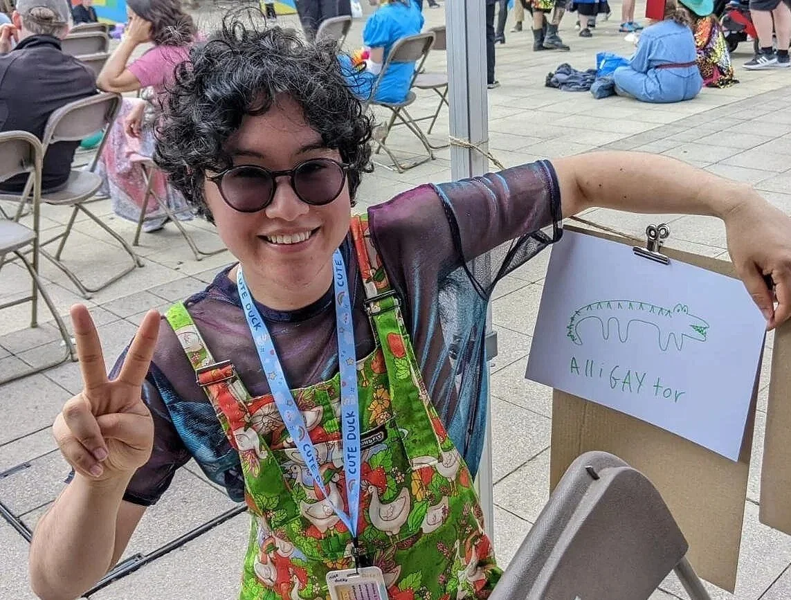 A person with curly hair, sunglasses, and a colorful apron is smiling and making a peace sign. They are holding a sign that says "ALL GAY for" with a drawing of a green dinosaur. The background shows people sitting at tables at an outdoor event or gathering.