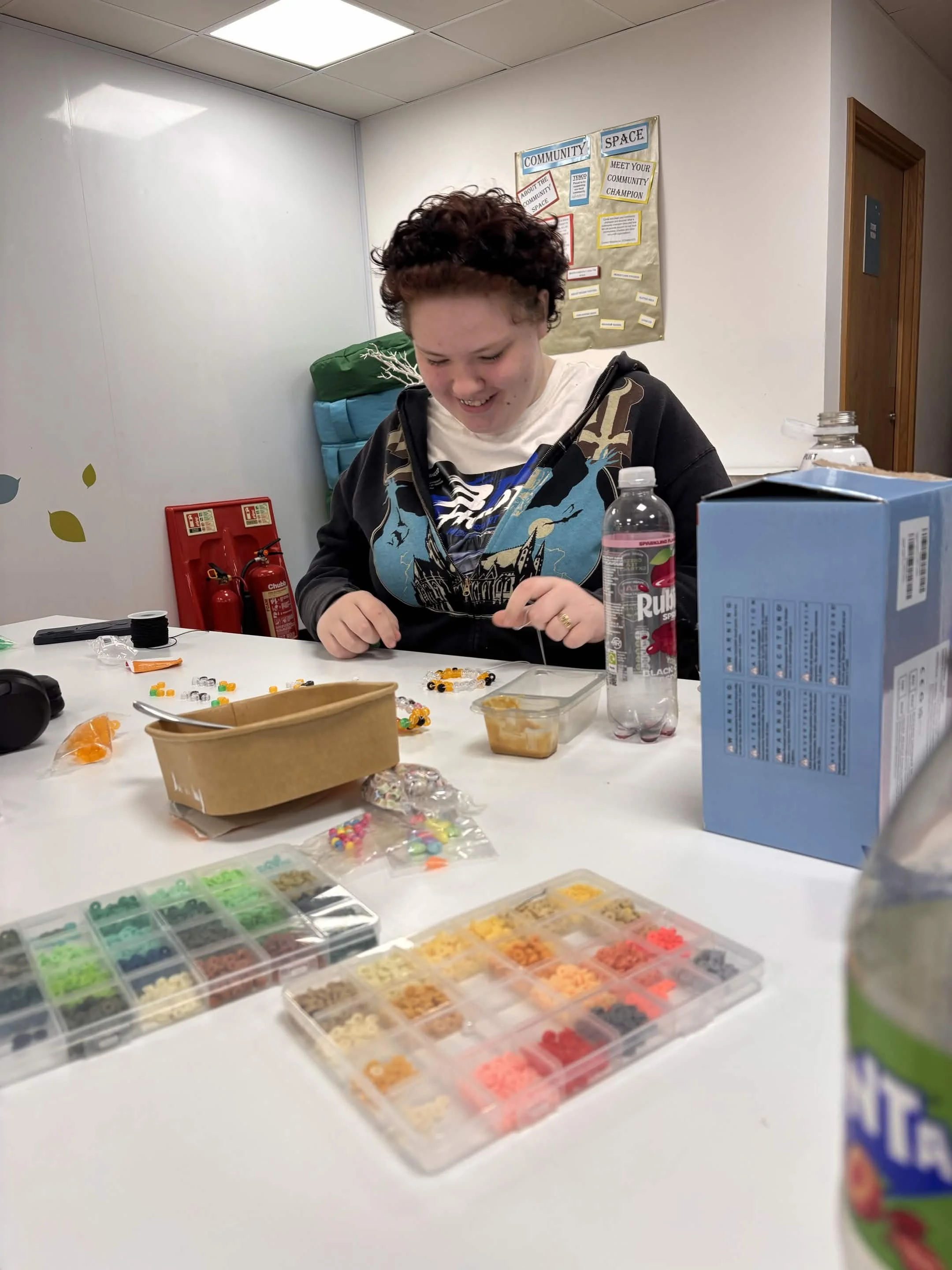 A person smiling and working on a beading project at a table with various beads and supplies, in a room with a bulletin board labeled 'Community Space' and a fire extinguisher in the background.