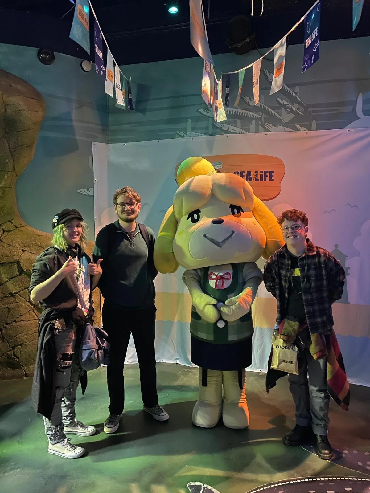 Three people and an animal cross character standing together.  The background has a banner with the word 'SEA LIFE' and hanging decorations.