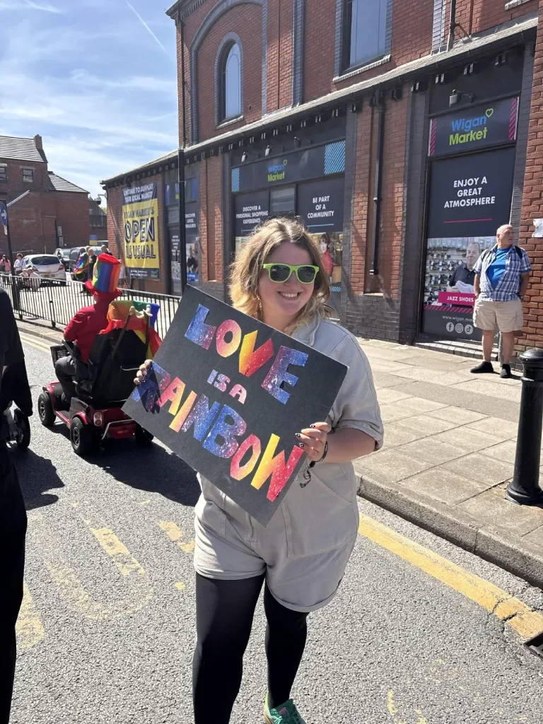 A person with green sunglasses holding a colorful sign that reads 'LOVE IS A RAINBOW' on a street during a sunny day.