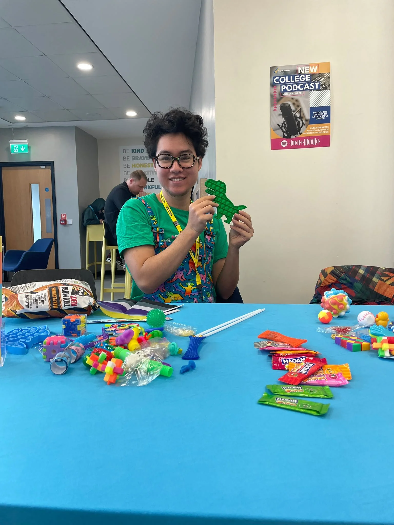 A person with short curly hair, glasses, and a lanyard is smiling and holding a green dinosaur-shaped toy at a table covered with colorful toys, candy, and small objects in an indoor space with chairs and walls decorated with motivational words and posters.