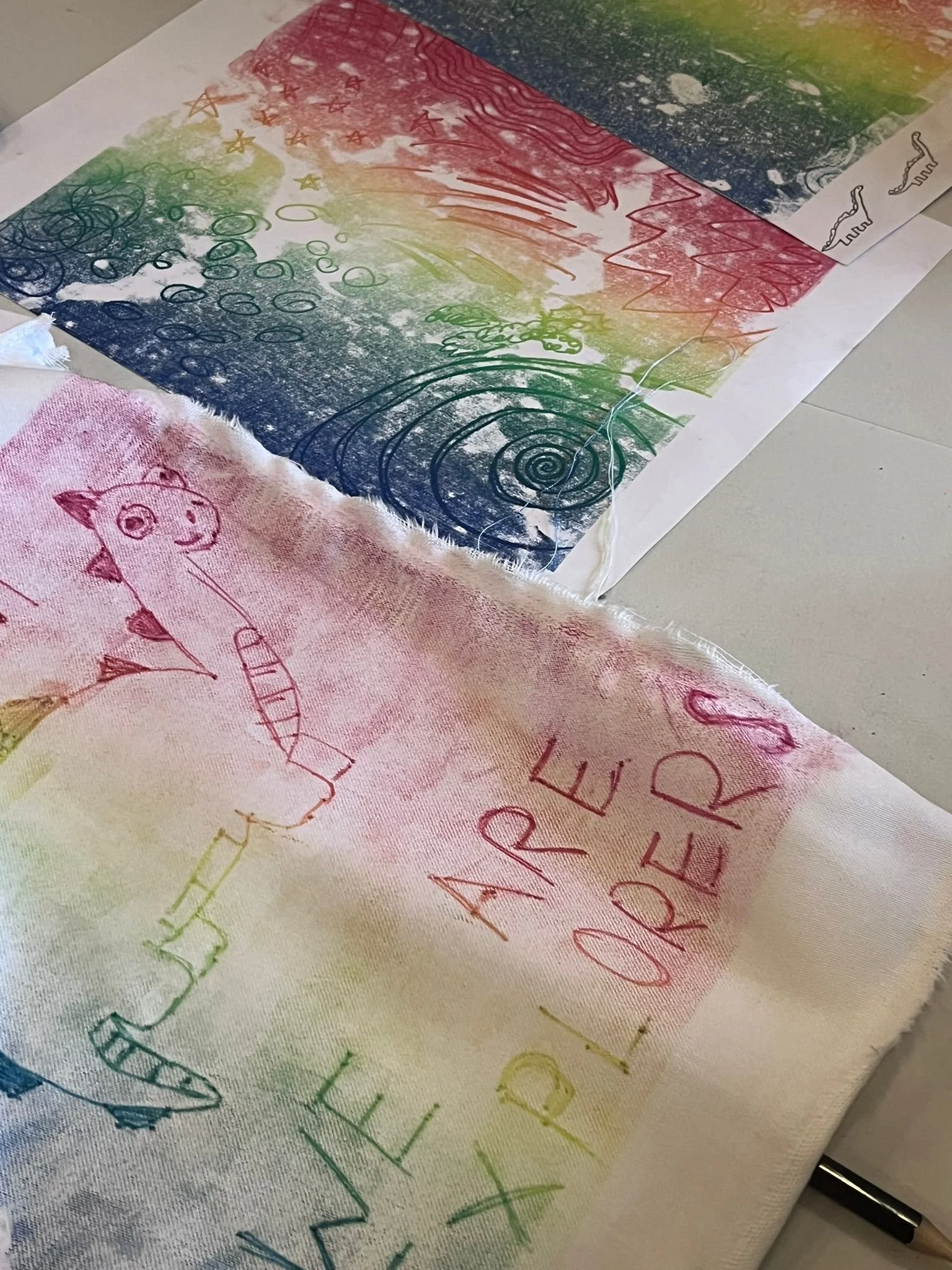 A piece of fabric with rainbow watercolor stripes and a rainbow-colored handwritten message that reads 'WE ARE EXPLORERS' in colorful letters, with a drawing of a dinosaur.
