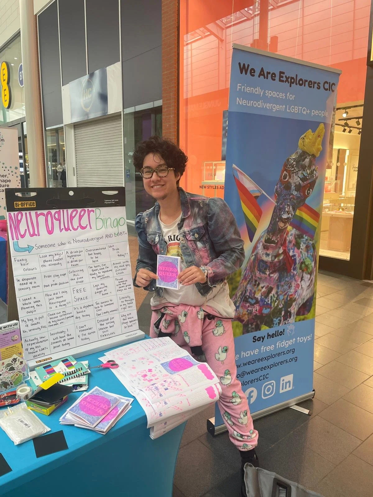 Person with short curly hair, glasses, and a big smile, wearing a denim jacket and pink pajamas with unicorn prints, standing at an informational booth about neurodiversity and LGBTQ+ inclusion, holding a flyer. The booth features a sign that says 'Neuro Queer Bingo' and a large colorful poster with a dog wearing rainbow accessories and a flag, with a background of orange and grey storefronts inside a mall.