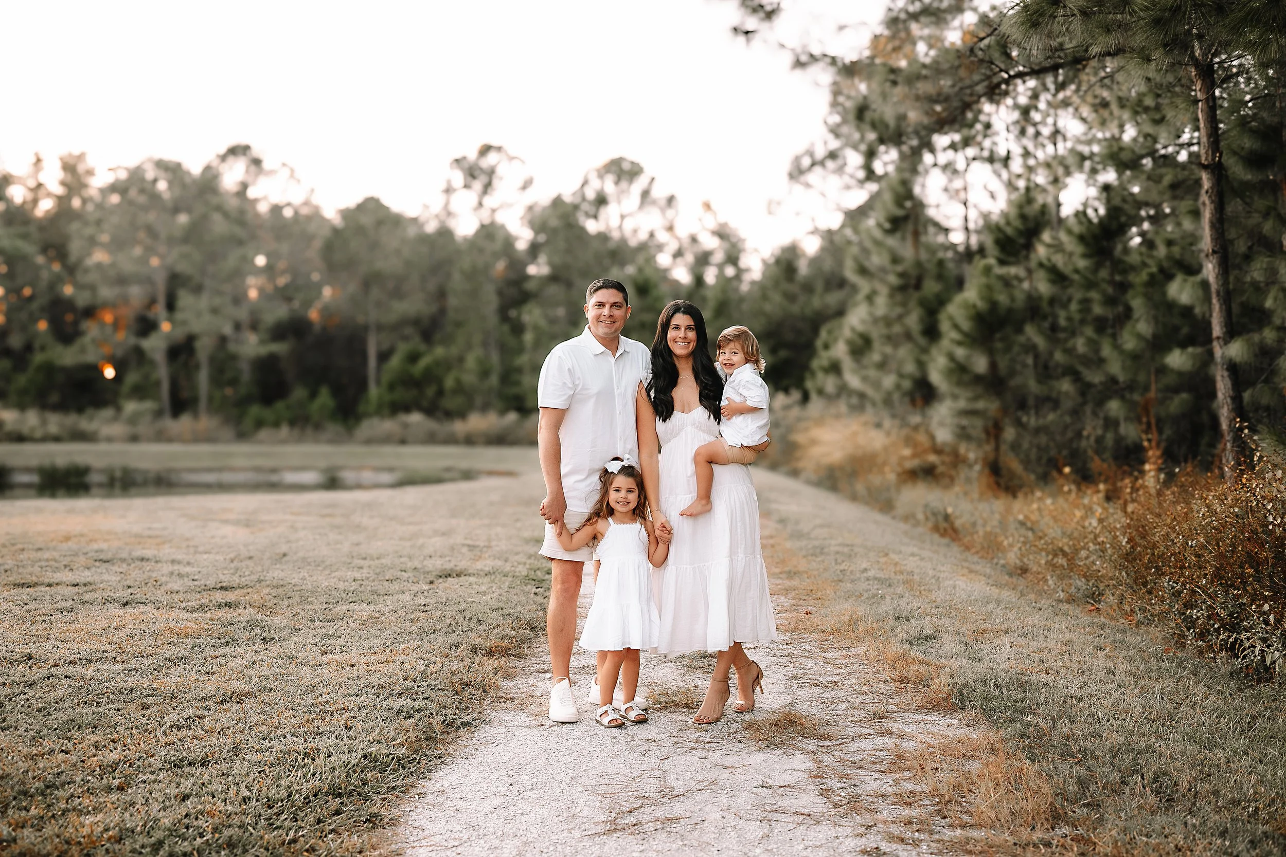 Family of four standing on a dirt path near a lake, with trees in the background, during sunset.