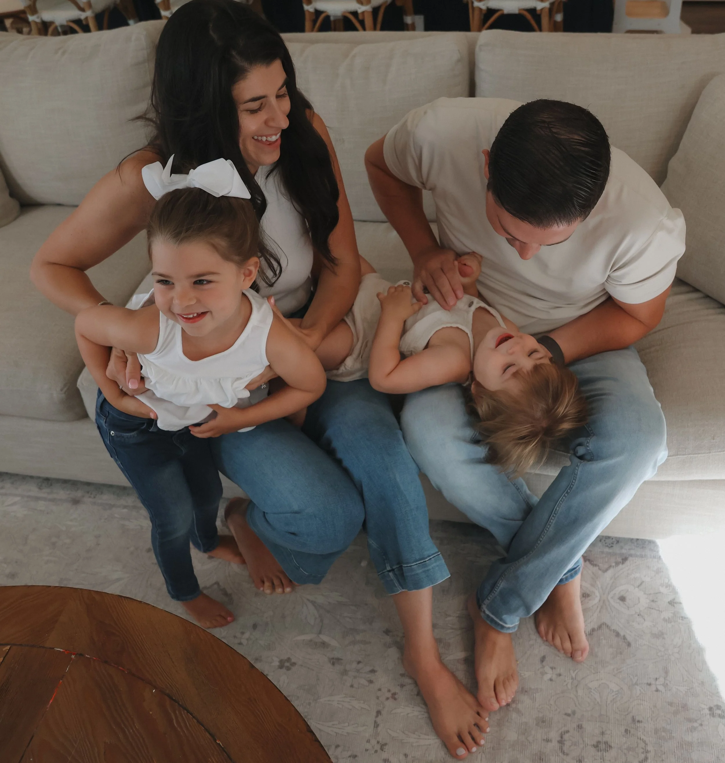 Family of four sitting on a beige sofa, playfully interacting with two young children. The girl with a white bow in her hair is smiling, one child is lying on a man's lap, laughing, while the woman is smiling and watching them, and the man is holding the other child, all barefoot in a cozy living room.