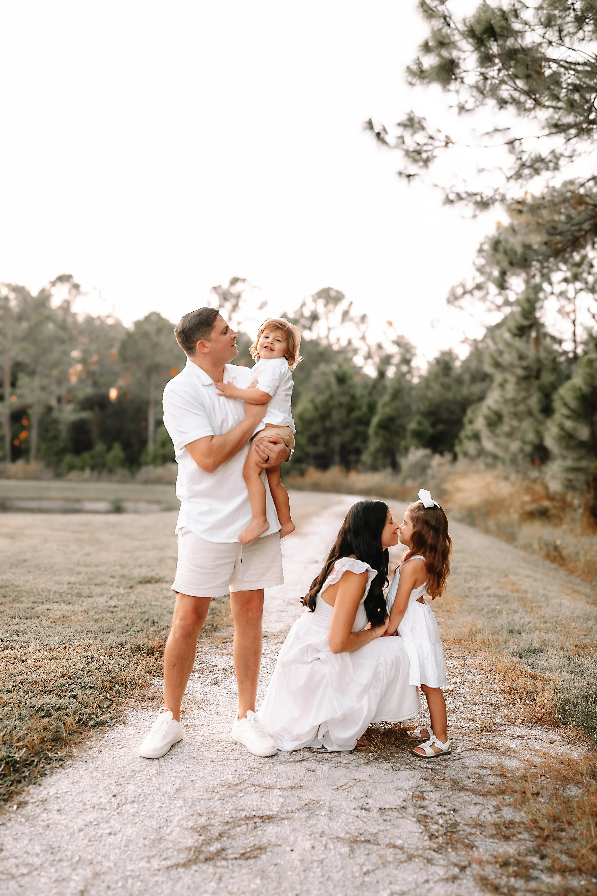 A family of four outdoors on a dirt path, with trees in the background. The father, dressed in a white shirt and beige shorts, holds a smiling young girl. The mother, kneeling and wearing a white dress, interacts with a young girl wearing a white dress and a bow, kissing her.