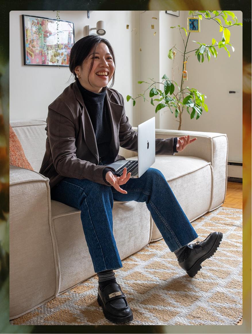 Therapist Phing Tan sitting on a beige sofa, holding a laptop, smiling, with a potted plant behind her and art on the wall.