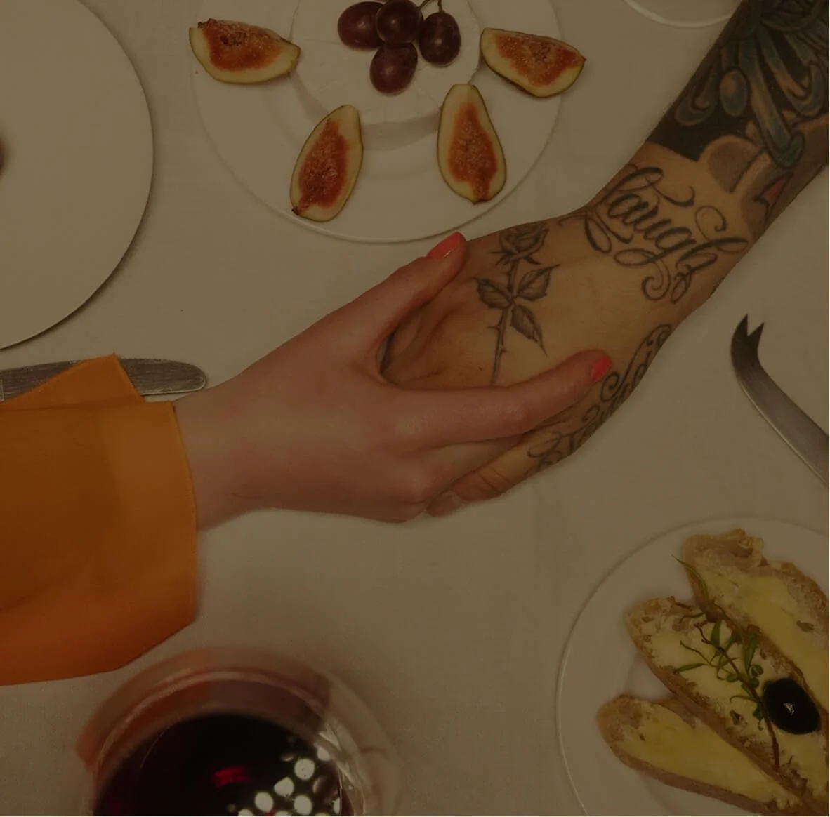 A tattooed arm being held by a person with a tan sleeve, at a dinner table with plates of figs, grapes, and assorted food. A glass of red wine is also visible.
