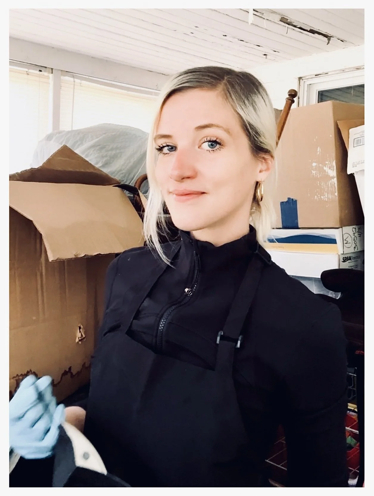 A woman with blonde hair and blue eyes, wearing a black jacket, is holding a syringe and preparing to give a vaccination or injection. She is in a room filled with cardboard boxes and supplies.