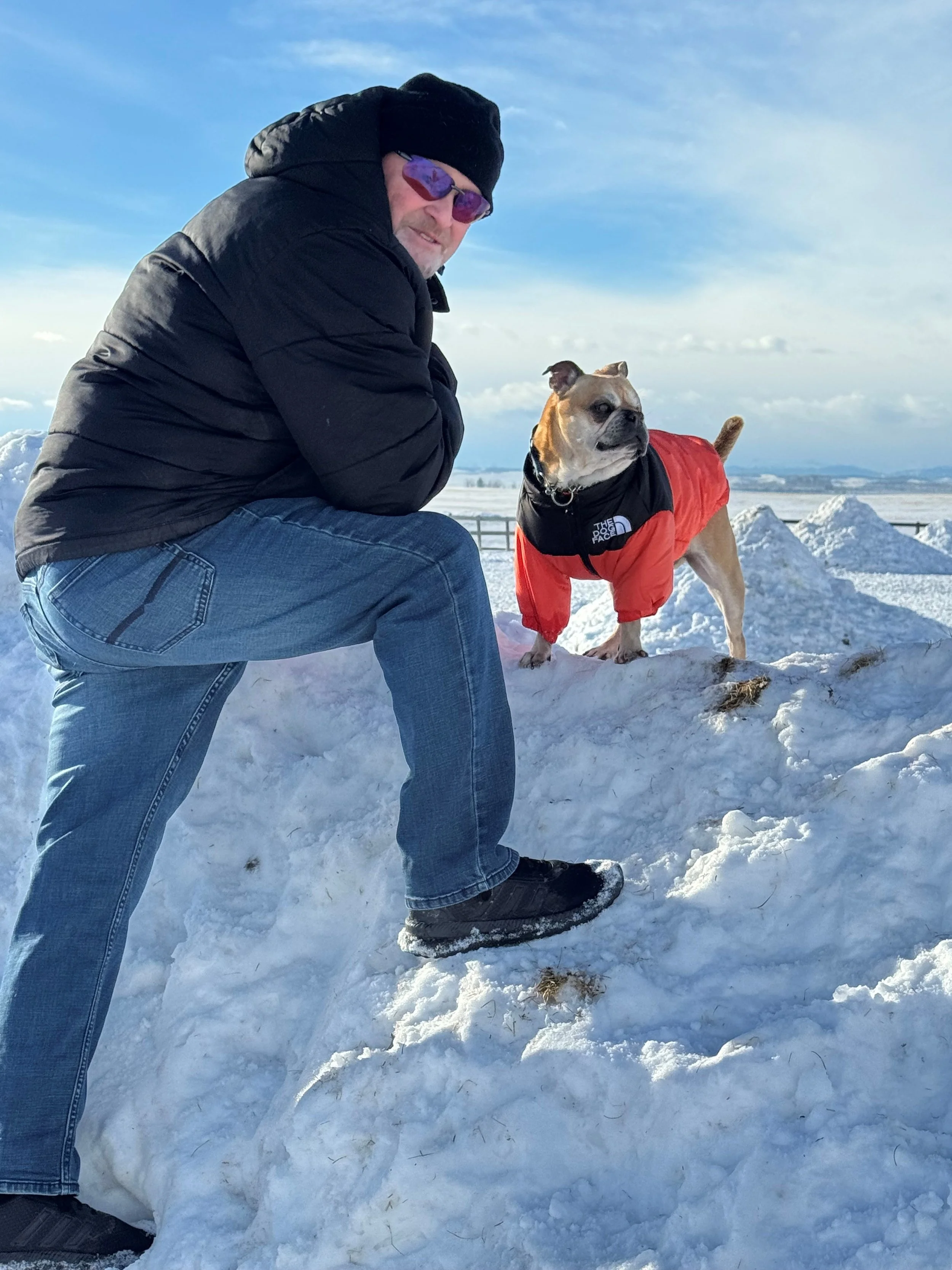 A man in a black jacket, jeans, and a black hat posing with a tan bulldog in a red and black jacket on snow-covered ground in a winter landscape.