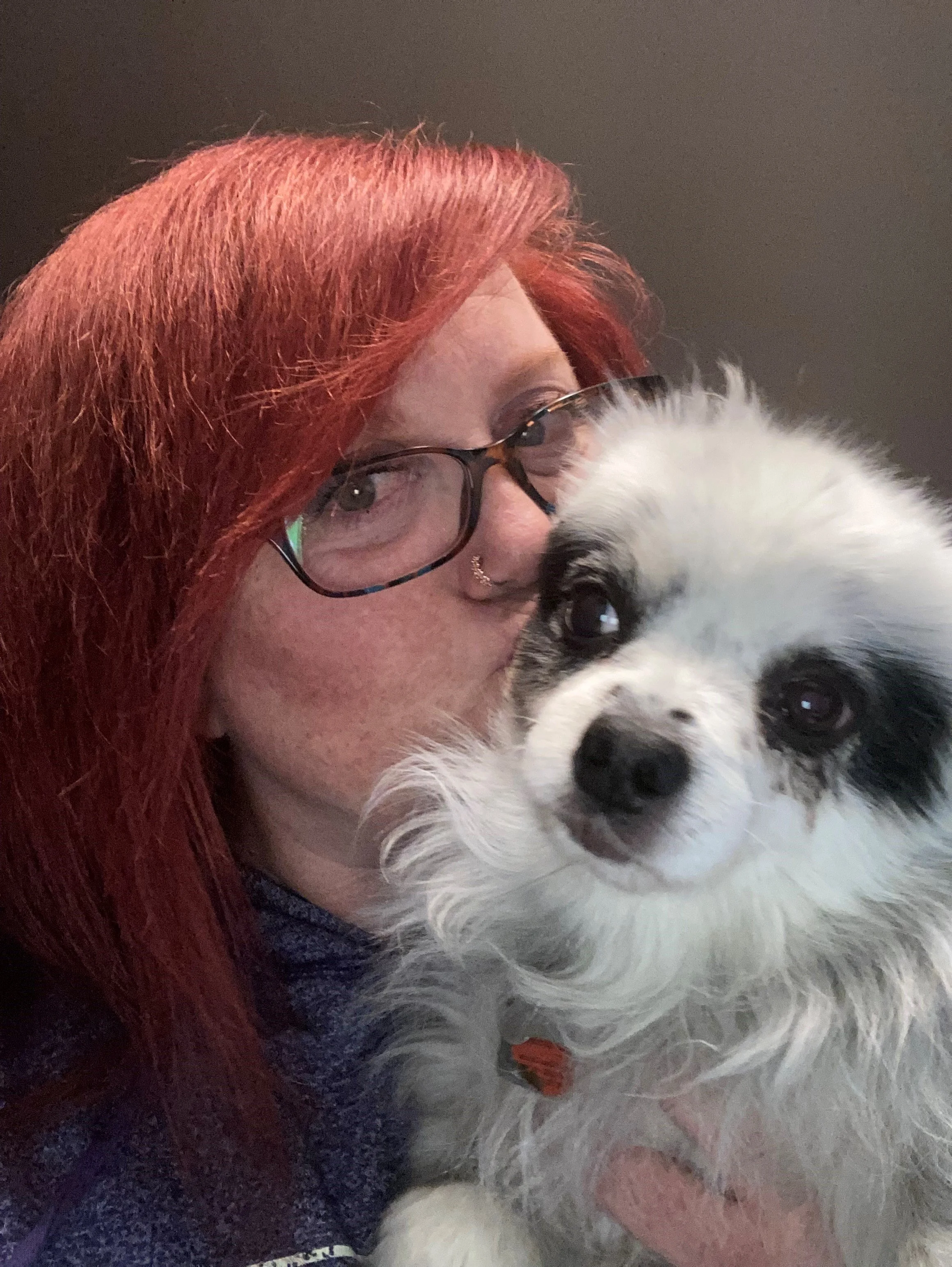 A woman with red hair and glasses holding a cute white dog with black markings close to her face.
