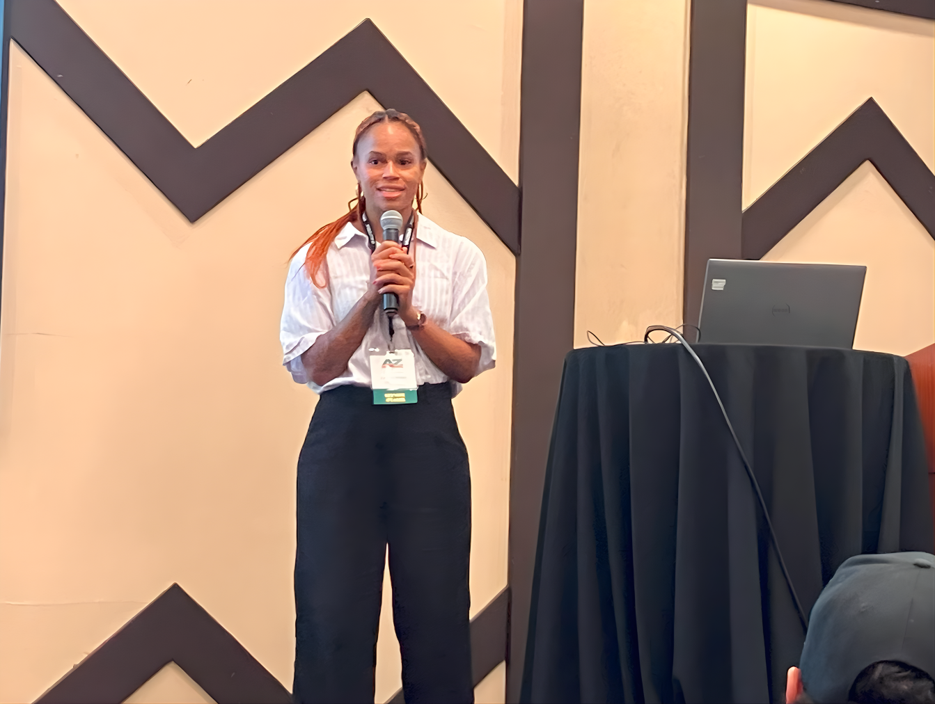 A woman stands on a stage holding a microphone, giving a presentation at a conference or seminar, with a computer on a small black table next to her.