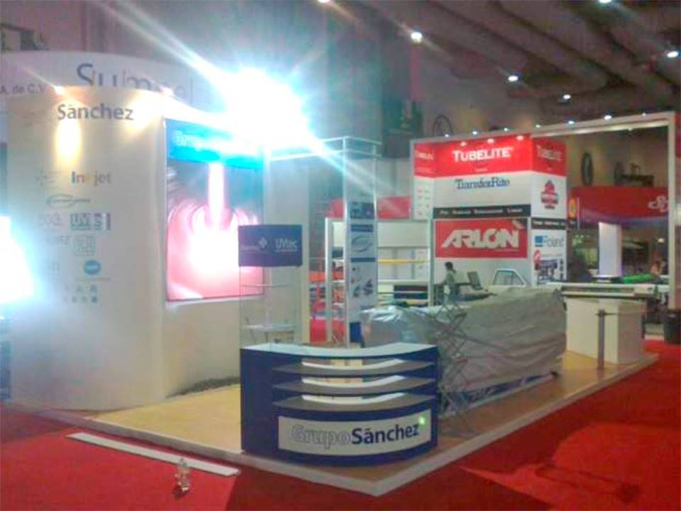 Imagen de un stand en una feria o exposición con el nombre 'Grupo Sánchez' en un cartel en la parte frontal. El stand tiene varios carteles y pantallas, algunos con marcas como Tubelite y Arlon. Hay mesas, sillas y materiales en el interior del stand.