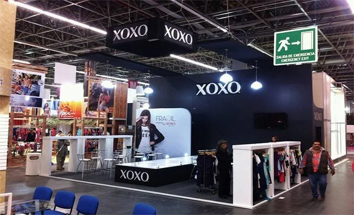 Stand de la marca XOXO en una feria o exposición, con ropa exhibida y una joven promocionando la marca, en un ambiente con señalización de salida de emergencia.