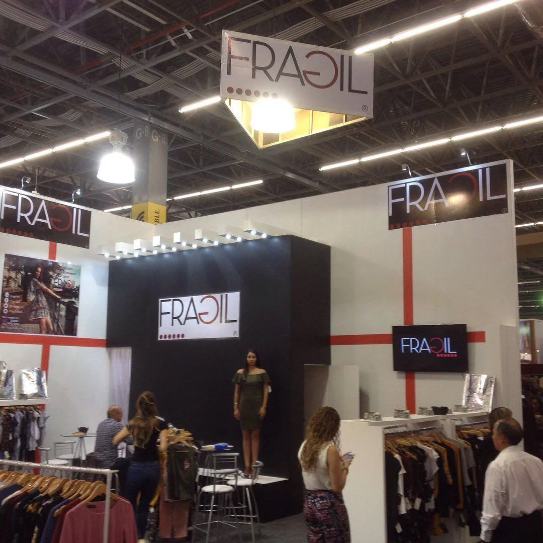 Stand de exhibición de ropa con señalización que dice FRAGIL, en un evento o feria comercial con varias personas interactuando y vestidas casualmente.