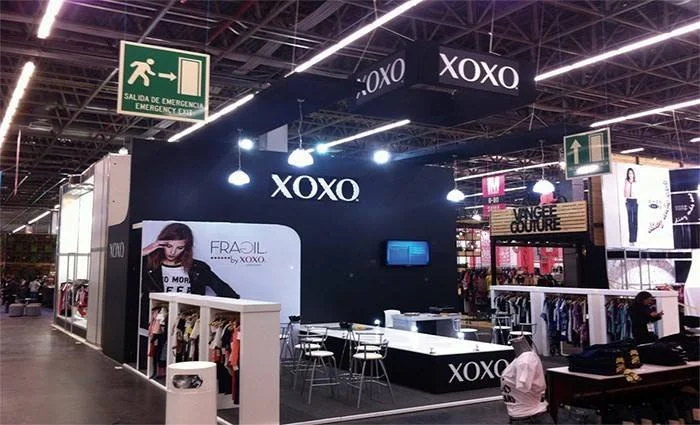 Pantalla de exhibición de la marca XOXO en una tienda dentro de una tienda, con sillas y mesas blancas, ropa exhibida y un cartel publicitario con una mujer.