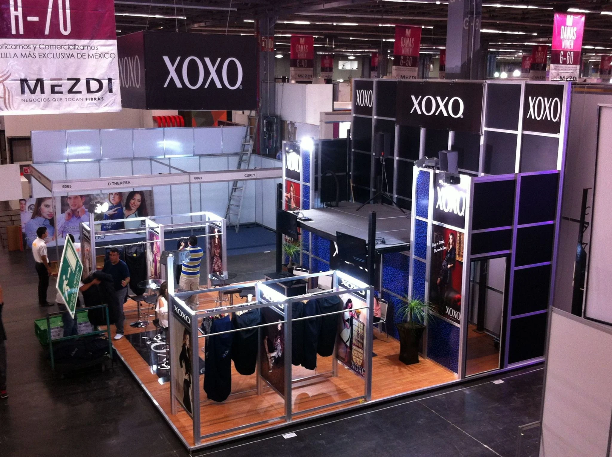 Estánd de exhibición en una feria con marcas XOXO y MEZDI, con una pequeña zona de degustación y una estructura para promociones, en un ambiente de exposición grande con personas y material de promoción.