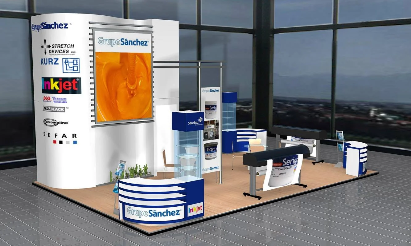 Estudio de exhibición con banner promocional, mesas, sillas y cubículos con logos de varias marcas como Grupo Sánchez, Injet, Serif, y otros, en una oficina con ventanas de gran tamaño.