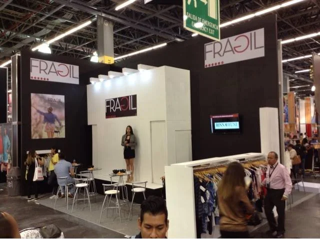 stand de exhibición de la marca 'FRAJIL' en una feria, con varias personas, sillas y ropa en exhibición.
