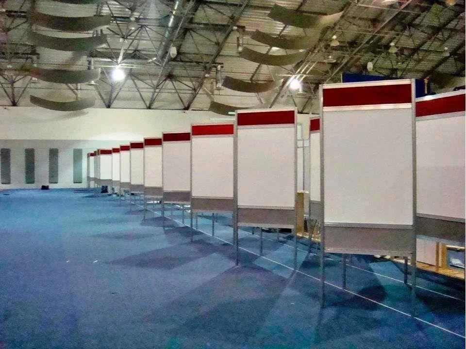 Filas de cabinas o stands vacíos en un espacio grande, posiblemente en un centro de convenciones o exposiciones, con techo alto y luces artificiales.