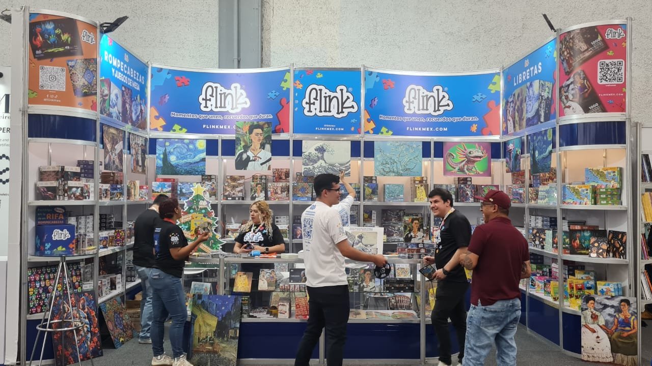 Pabellón de exhibición de la editorial Flink con estantes llenos de libros, un árbol de Navidad decorado, varias personas comprando y conversando, carteles con arte y promociones, y banners azules en el fondo.
