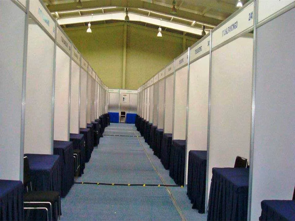 Pasillos de una feria o exposición con cabinas o stands vacíos y sillas cubiertas con manteles oscuros a los lados.